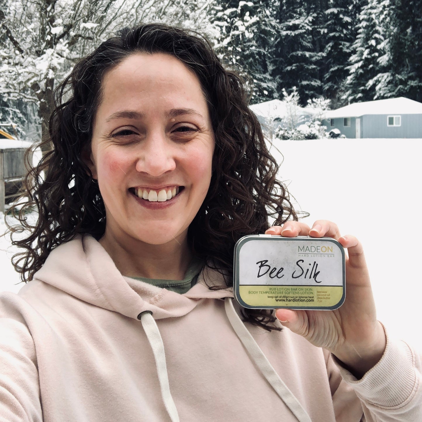 curly-haired, smiling woman in the snow holding a beeswax-based beesilk lotion bar for dry, winter skin